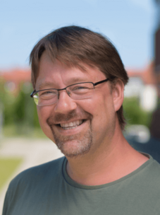 Herr Szameit mit Brille und Bart, draußen im Sonnenlicht lächelnd.